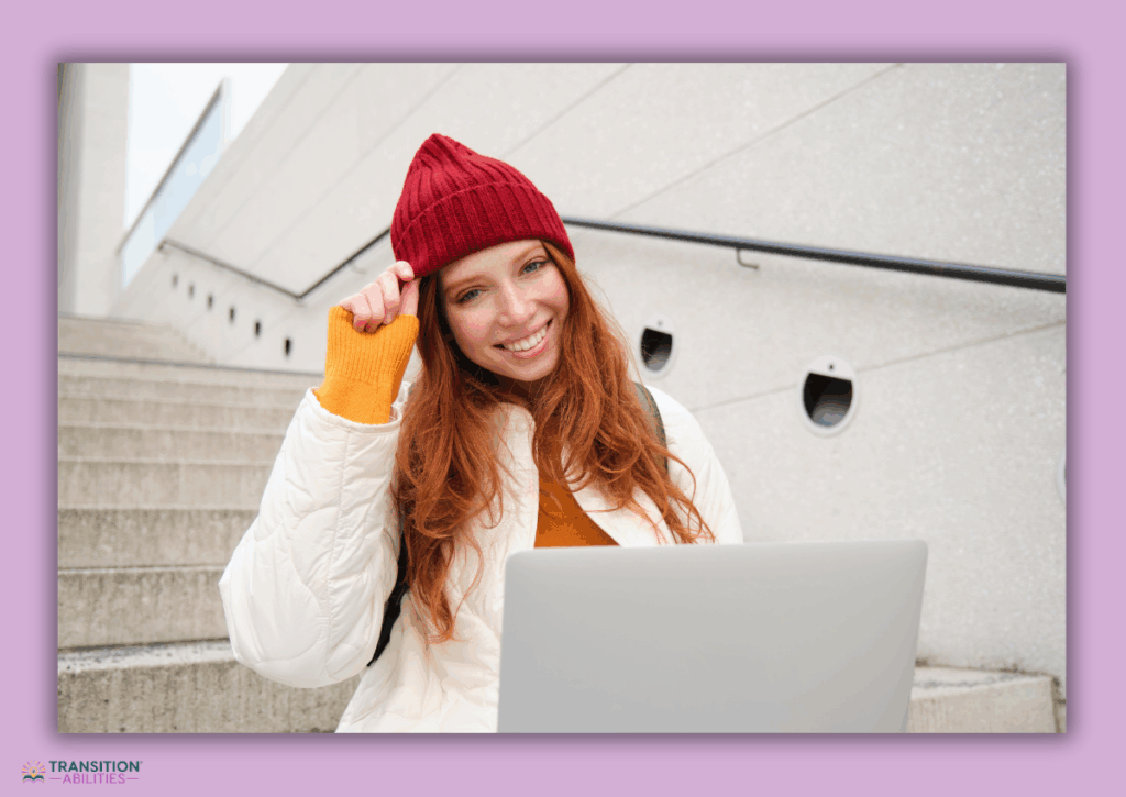 Smiling student wearing a red hat and winter coat using a laptop outdoors.