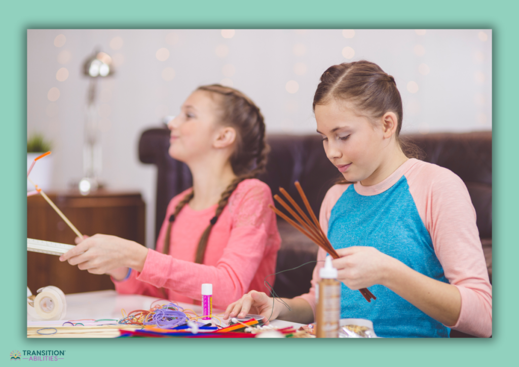 Two girls at a table focused on a craft project using colorful sticks, glue, and other art supplies.