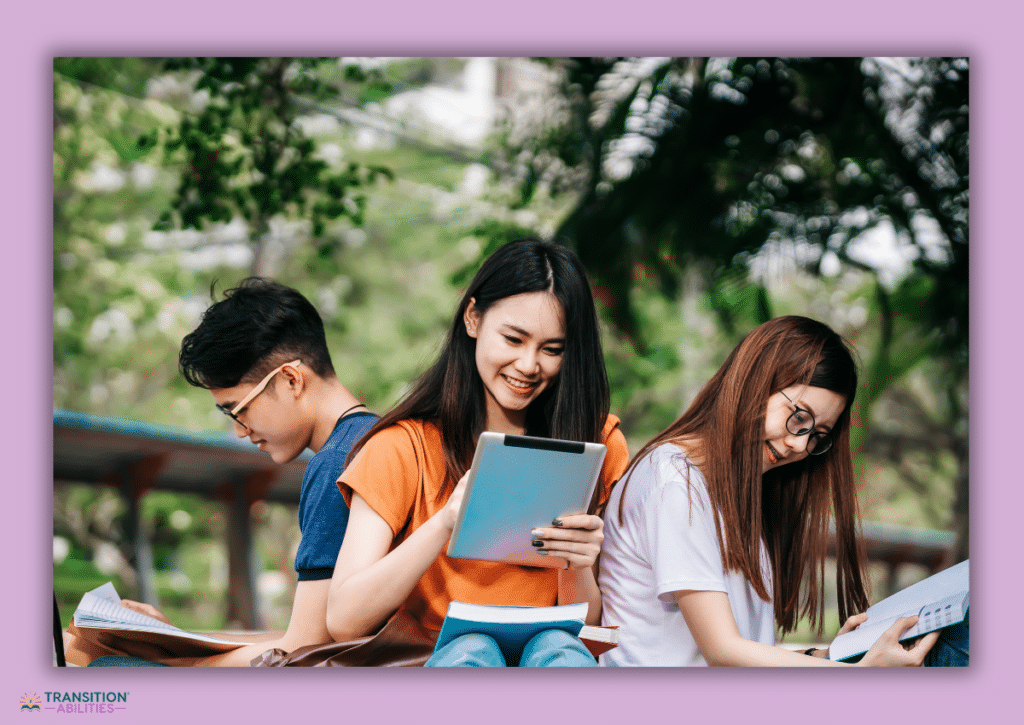 Three high school students sit outside, smiling while using tablets and reading books.