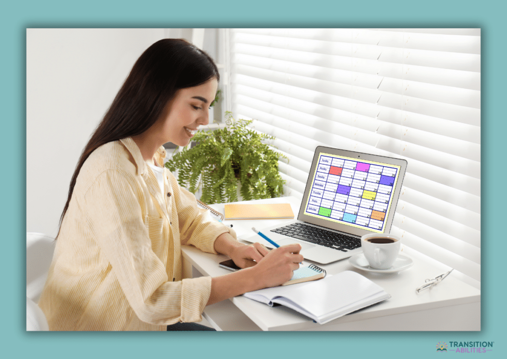 A young woman writes in a notebook at a desk while looking at a colorful visual schedule displayed on her laptop.