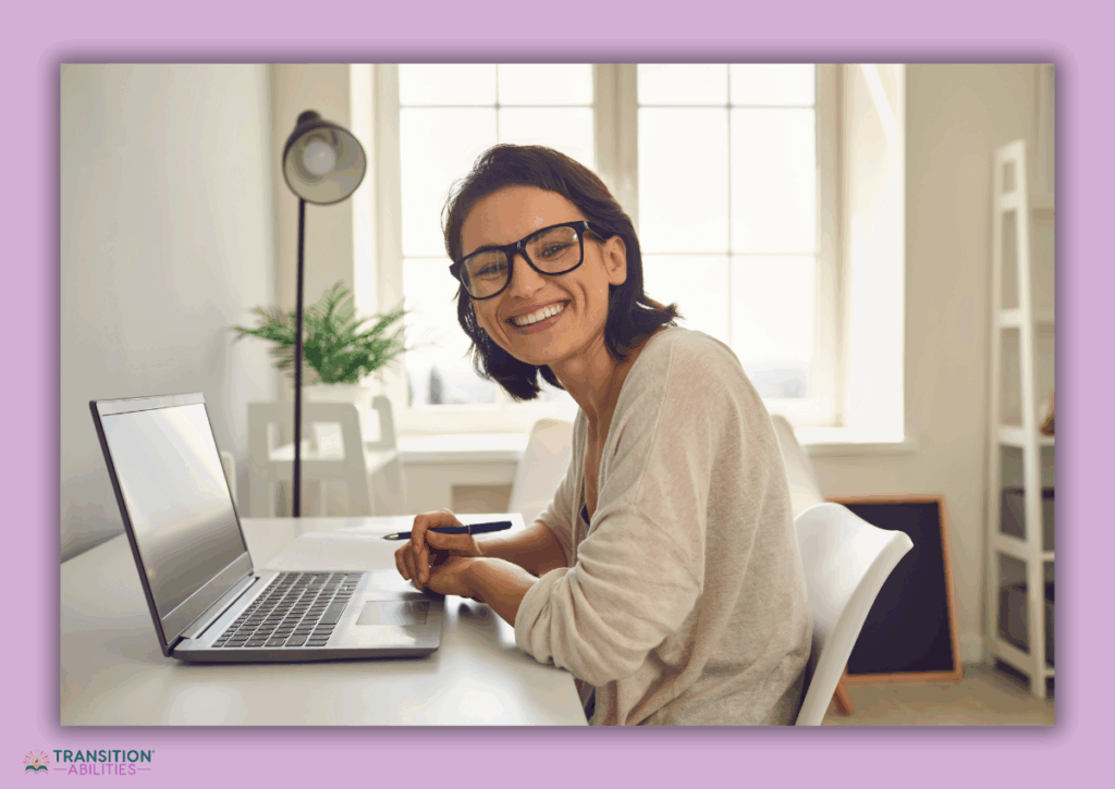 Smiling woman with glasses sitting at a desk with a laptop, holding a pen in a bright home office