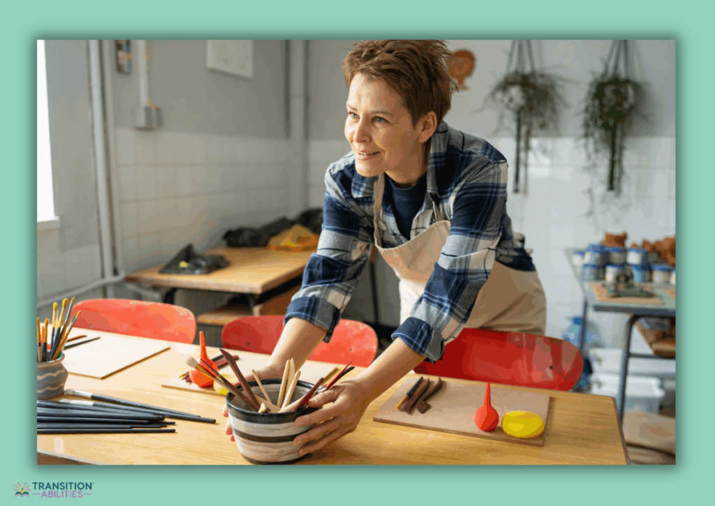 A woman smiles while organizing pottery tools on a table in an art studio.