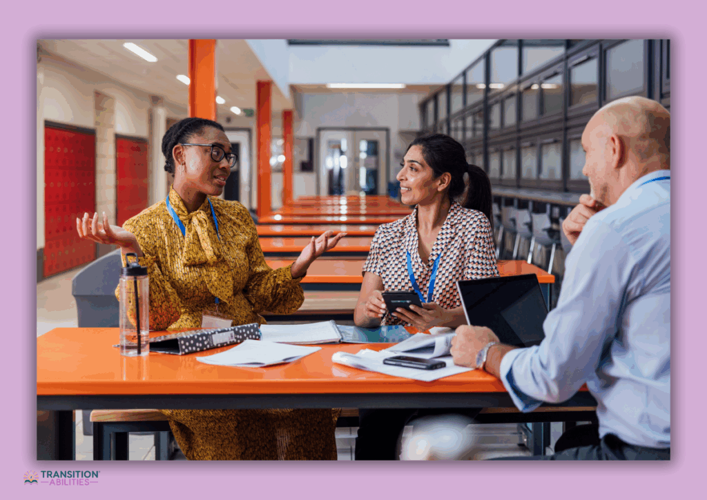 Three school staff members having a discussion at a cafeteria table with notebooks and devices