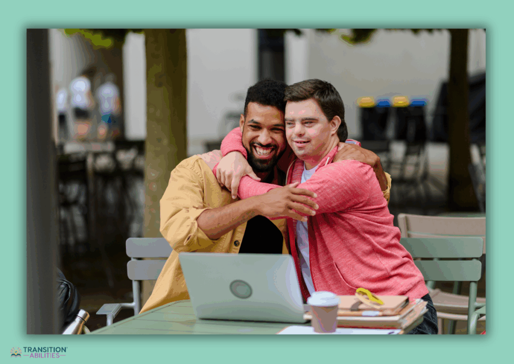 Two men smiling and hugging while sitting at an outdoor table with a laptop.